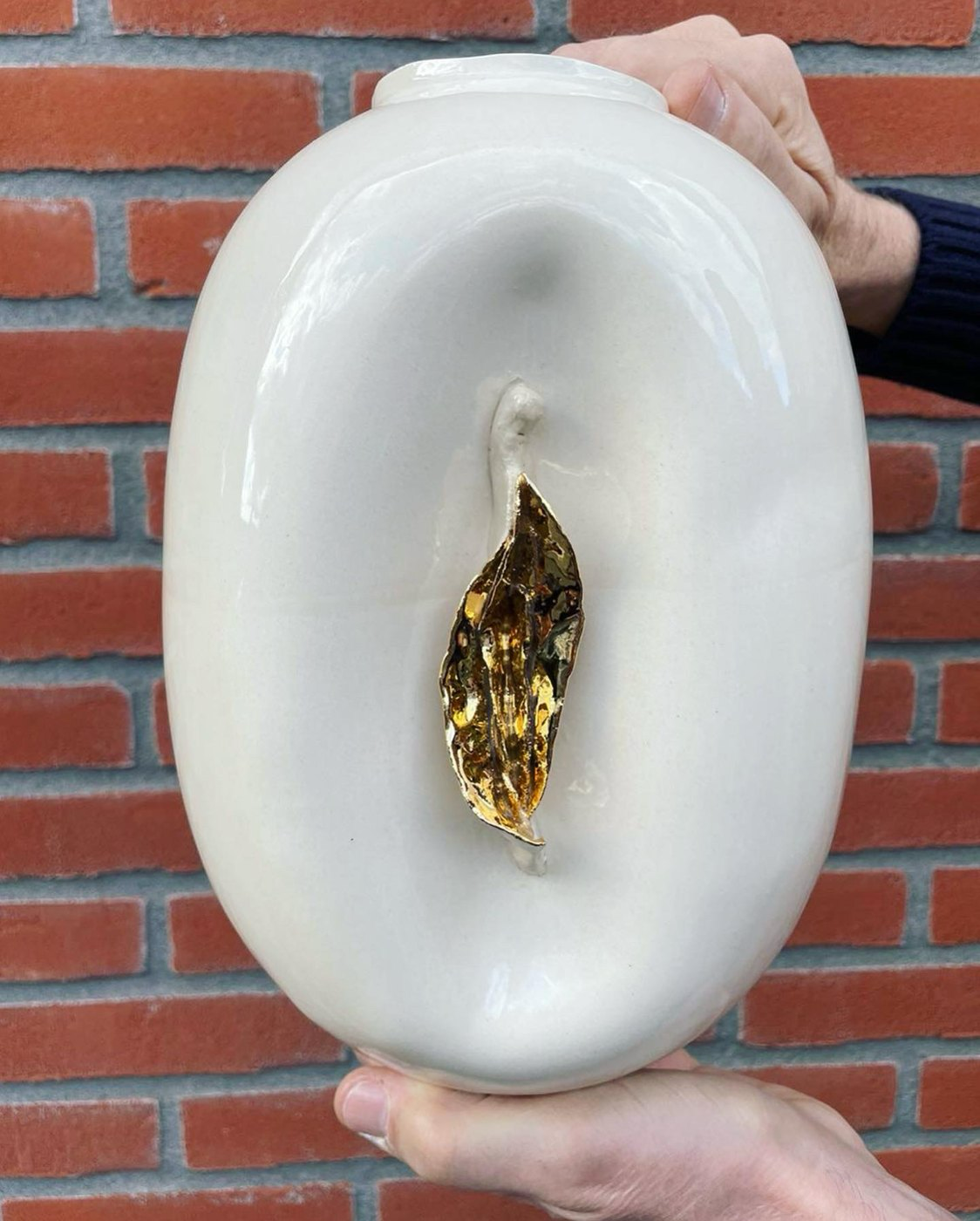 White ceramic Vulvase with a gold detail held in front of a brick wall